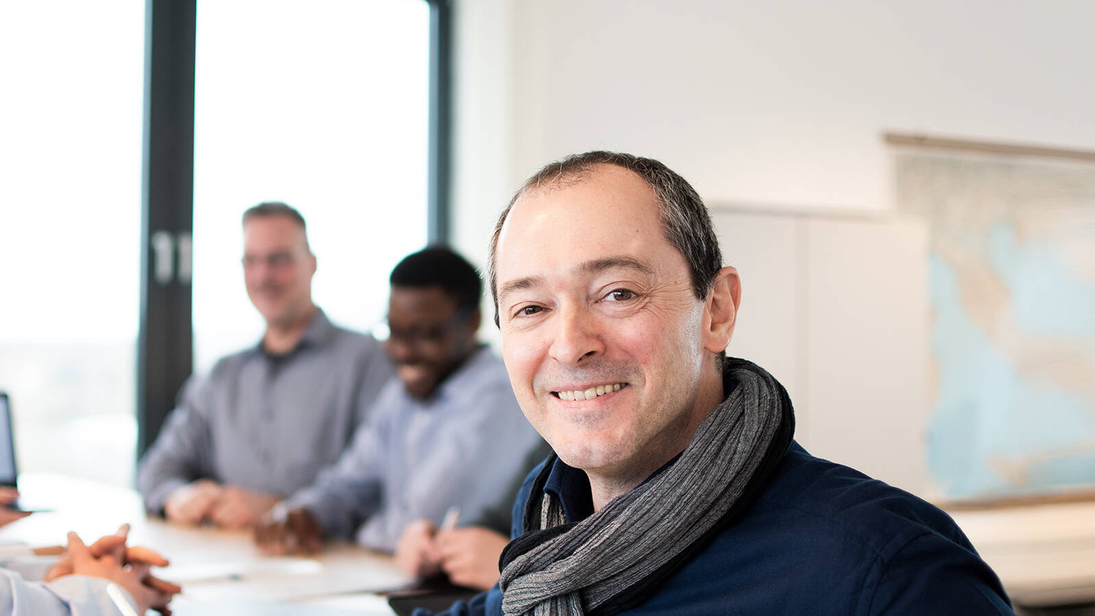 A man stands at a meeting table with several people, smiling at the camera