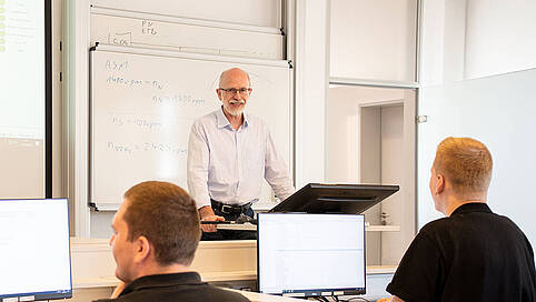 A trainer stands at a lectern in front of a whiteboard and explains