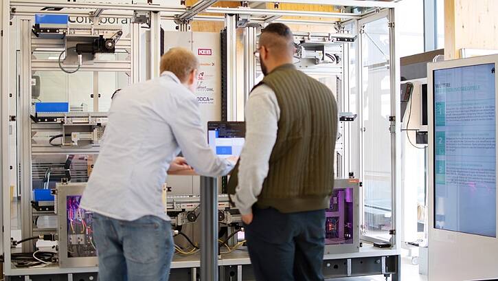 two men standing and programming on a laptop in front of an eletrotechnical setup