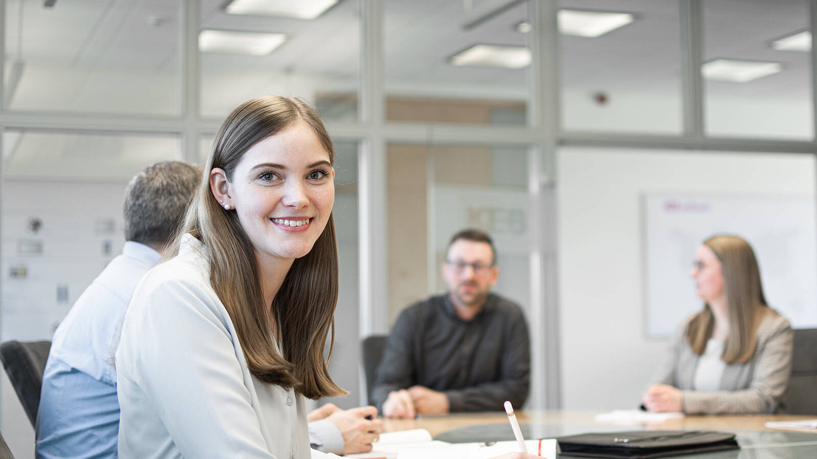 A woman sits at a meeting table with several people, smiling at the camera