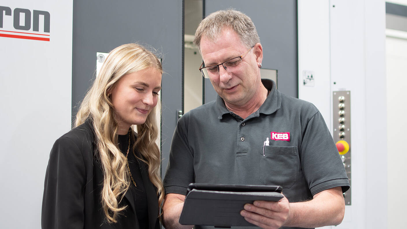 A woman and a man stand in front of a machine and look into a tablet together