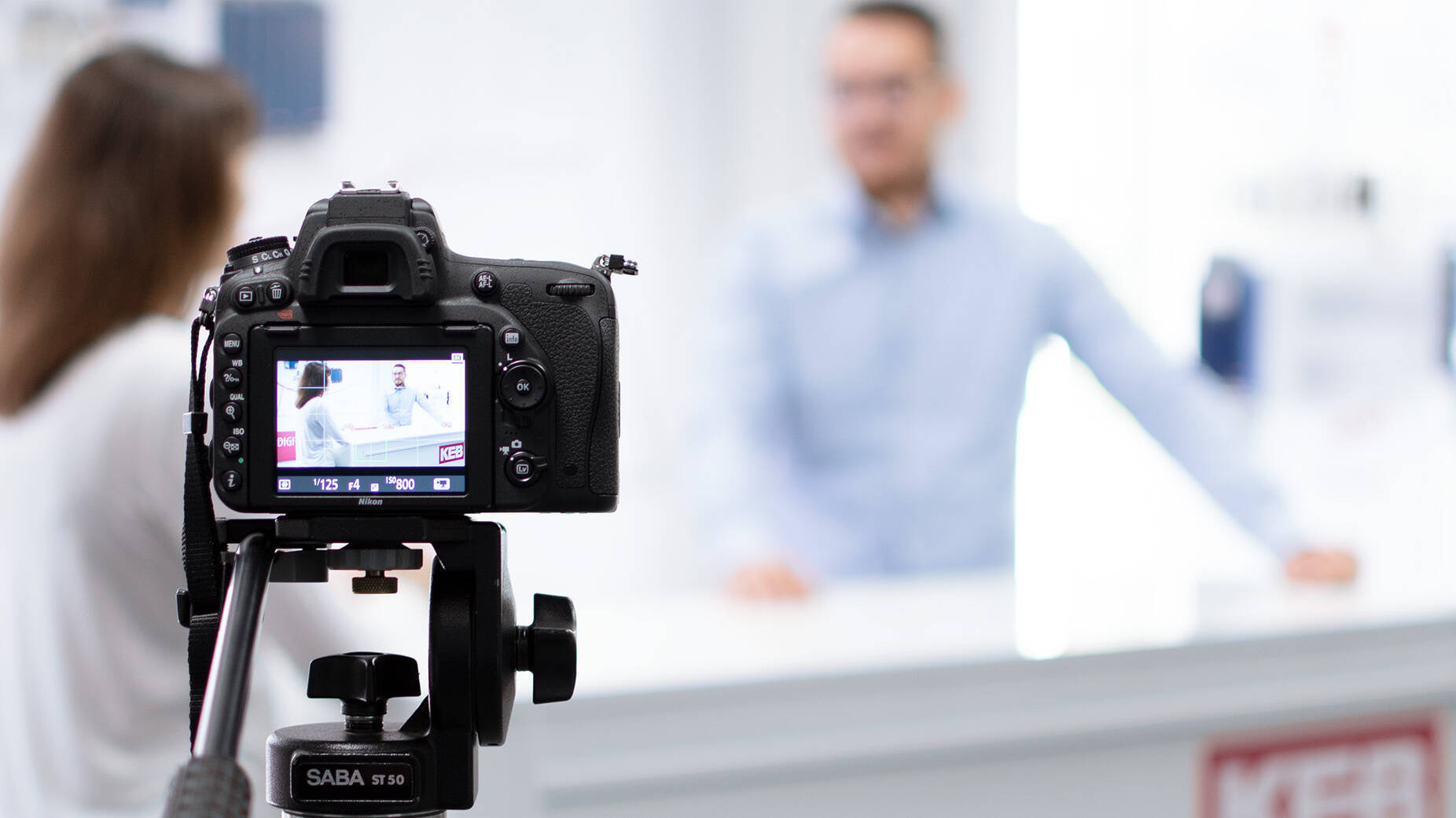 a camera in the foreground, filming to persons at a desk in the background doing a webinar