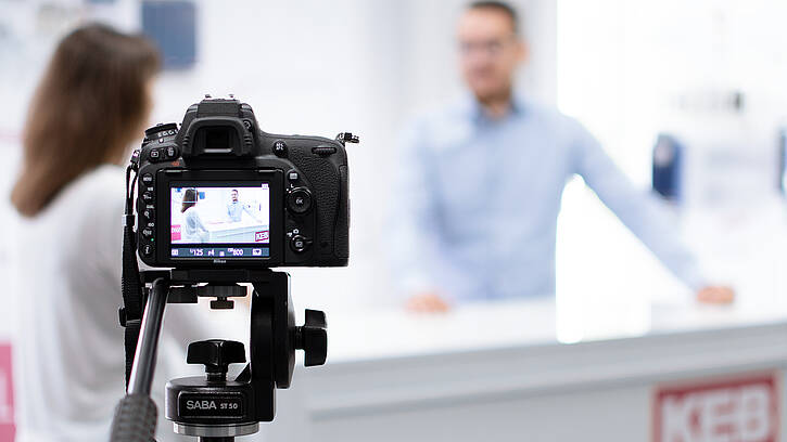 a camera in the foreground, filming to persons at a desk in the background doing a webinar