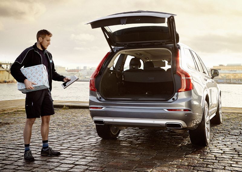 Man with package and tablet stands next to an SUV with an open trunk