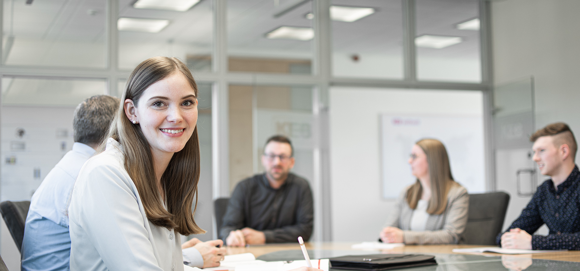 A woman sits at a meeting table with several people, smiling at the camera