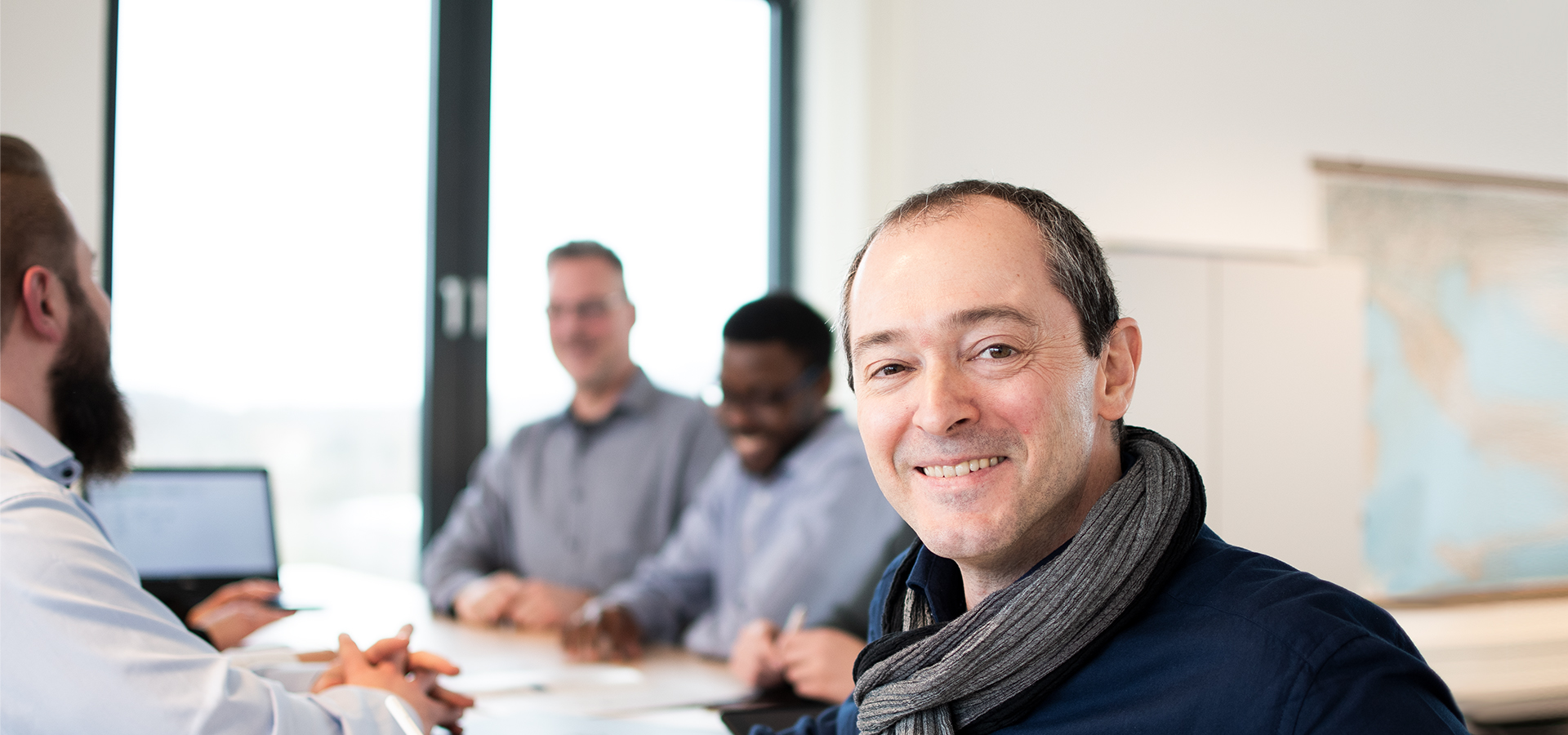 A man sits at a meeting table with several people, smiling at the camera