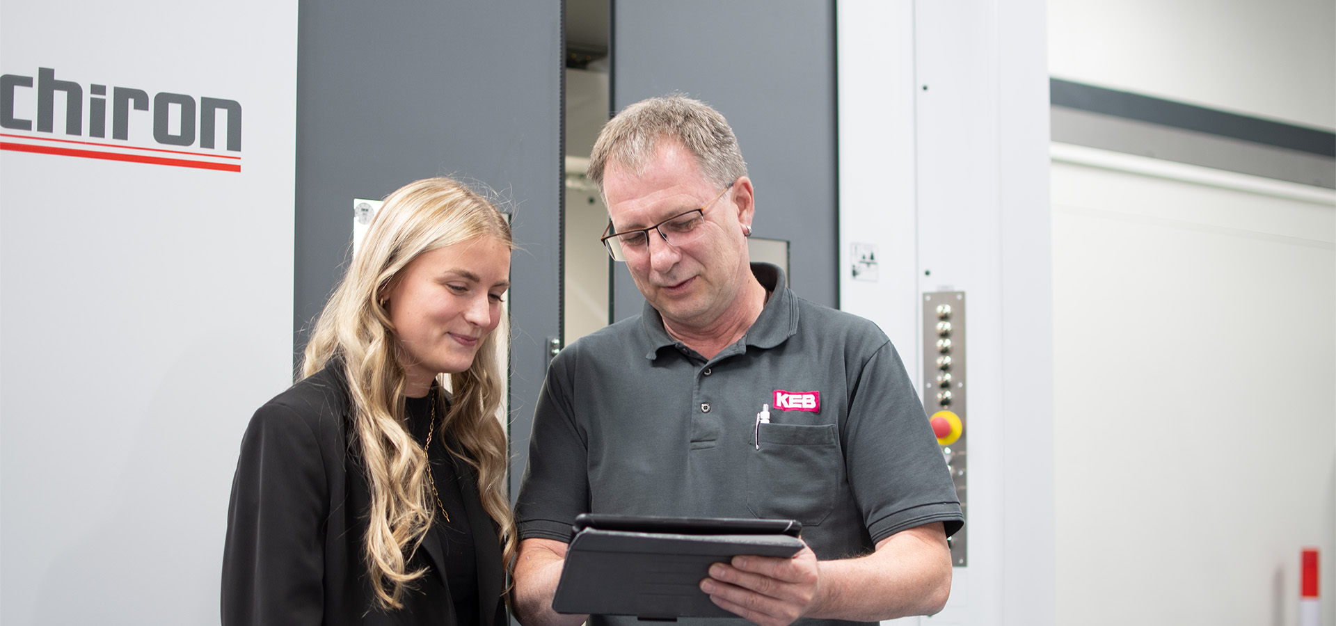 A woman and a man stand in front of a machine and look into a tablet together